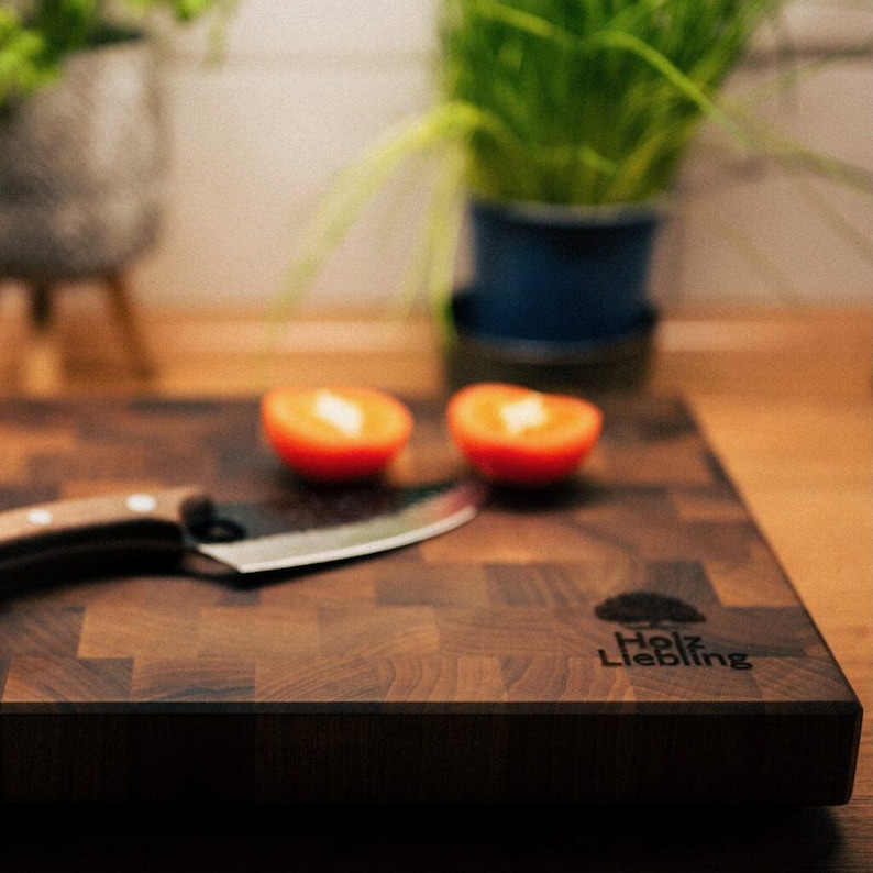 May include: A dark brown end-grain wooden cutting board with a checkerboard pattern. The board features a black logo of a tree and the text "Holz Liebling" branded on the surface. A metal knife and two halved orange tomatoes rest on the board.