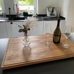 May include: A wooden cutting board with a natural wood finish and a raised edge. Two wine glasses and a bottle of champagne are on the board. A vase with dried flowers is in the background.