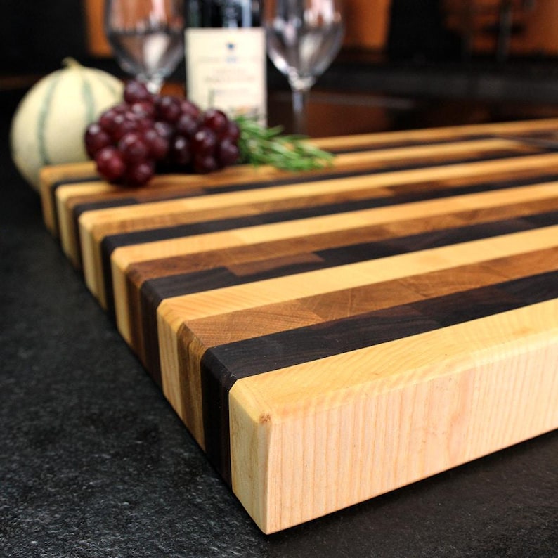 Peut inclure: Une planche &agrave; d&eacute;couper rectangulaire en bois pr&eacute;sentant des bandes horizontales altern&eacute;es de bois d'&eacute;rable clair et de bois de noyer fonc&eacute;. La planche a une construction &eacute;paisse et solide avec une surface lisse finie montrant le grain naturel.