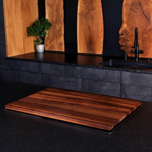 May include: A rectangular wooden chopping board with a rich, dark brown grain pattern and smooth edges. The board is positioned horizontally on a dark, textured surface, showcasing a natural wood finish with visible linear wood grain details.