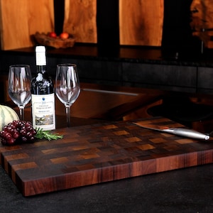 May include: A rectangular wooden chopping board with a dark brown end-grain checkerboard pattern. Beside it sits a bottle of Chateau Roquelourbe red wine, two clear glass wine glasses, a cluster of red grapes, a sprig of rosemary, and a metal knife.