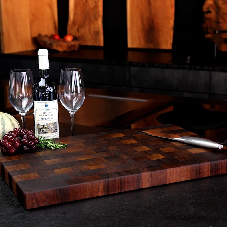 May include: A rectangular wooden chopping board with a dark brown end-grain checkerboard pattern. Beside it sits a bottle of Chateau Roquelourbe red wine, two clear glass wine glasses, a cluster of red grapes, a sprig of rosemary, and a metal knife.