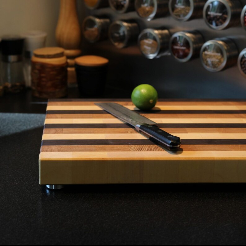 Peut inclure: Une planche &agrave; d&eacute;couper rectangulaire en bois pr&eacute;sentant des rayures horizontales altern&eacute;es de bois clair et fonc&eacute;. Un couteau de chef en acier inoxydable &agrave; manche noir repose en diagonale sur la surface &agrave; c&ocirc;t&eacute; d'un citron vert entier.