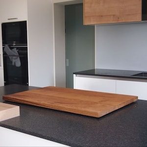 May include: A large, rectangular wooden cutting board sits on a black countertop in a kitchen. The board is made of light brown wood with a natural grain pattern.