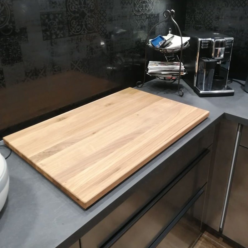 May include: A large, rectangular, light brown wooden cutting board sits on a gray countertop. The countertop has two drawers with black handles. The cutting board is made of wood with a natural grain pattern.