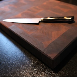 May include: A rectangular end-grain wooden chopping board featuring a dark brown checkerboard pattern with a smooth finish. A stainless steel kitchen knife with a black handle and two metallic rivets rests horizontally across the board surface.