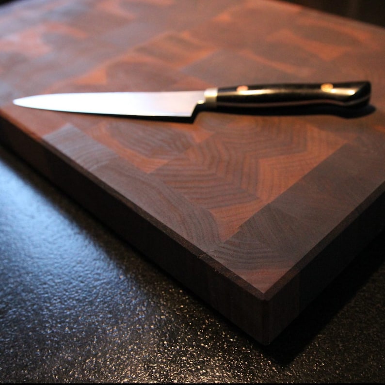 May include: A rectangular end-grain wooden chopping board featuring a dark brown checkerboard pattern with a smooth finish. A stainless steel kitchen knife with a black handle and two metallic rivets rests horizontally across the board surface.