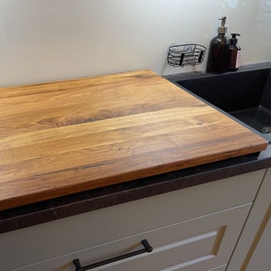 May include: A large, rectangular wooden cutting board sits on a black countertop next to a kitchen sink. The cutting board has a natural wood grain pattern and is made of light brown wood.