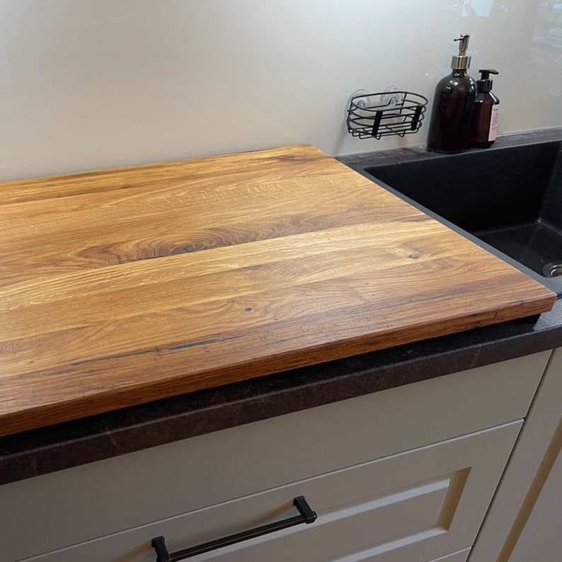 May include: A large, rectangular wooden cutting board sits on a black countertop next to a kitchen sink. The cutting board has a natural wood grain pattern and is made of light brown wood.