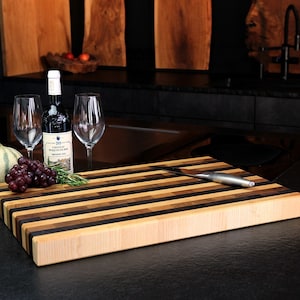 Peut inclure: Une planche &agrave; d&eacute;couper rectangulaire en bois compos&eacute;e de bandes horizontales altern&eacute;es de bois d'&eacute;rable clair, de noyer moyen et de weng&eacute; fonc&eacute;. Un couteau en acier inoxydable repose sur la planche, avec une bouteille de vin rouge, deux verres et du raisin.