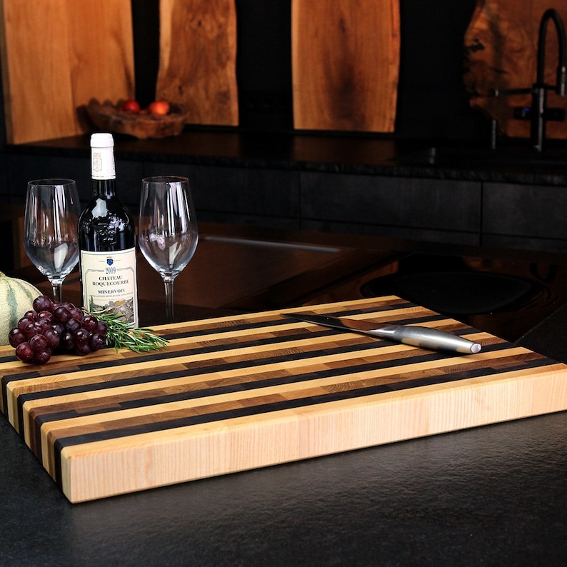 Peut inclure: Une planche &agrave; d&eacute;couper rectangulaire en bois compos&eacute;e de bandes horizontales altern&eacute;es de bois d'&eacute;rable clair, de noyer moyen et de weng&eacute; fonc&eacute;. Un couteau en acier inoxydable repose sur la planche, avec une bouteille de vin rouge, deux verres et du raisin.