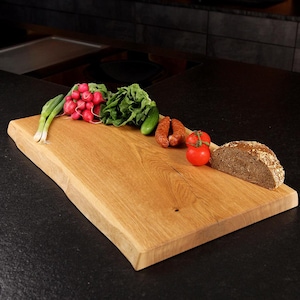 Può includere: Un tagliere rettangolare in legno con bordo naturale e venature a vista. Sulla superficie sono disposti cipollotti, un mazzetto di ravanelli rossi, foglie verdi, un cetriolo, due salsicce, due pomodori rossi e una fetta di pane ai cereali.