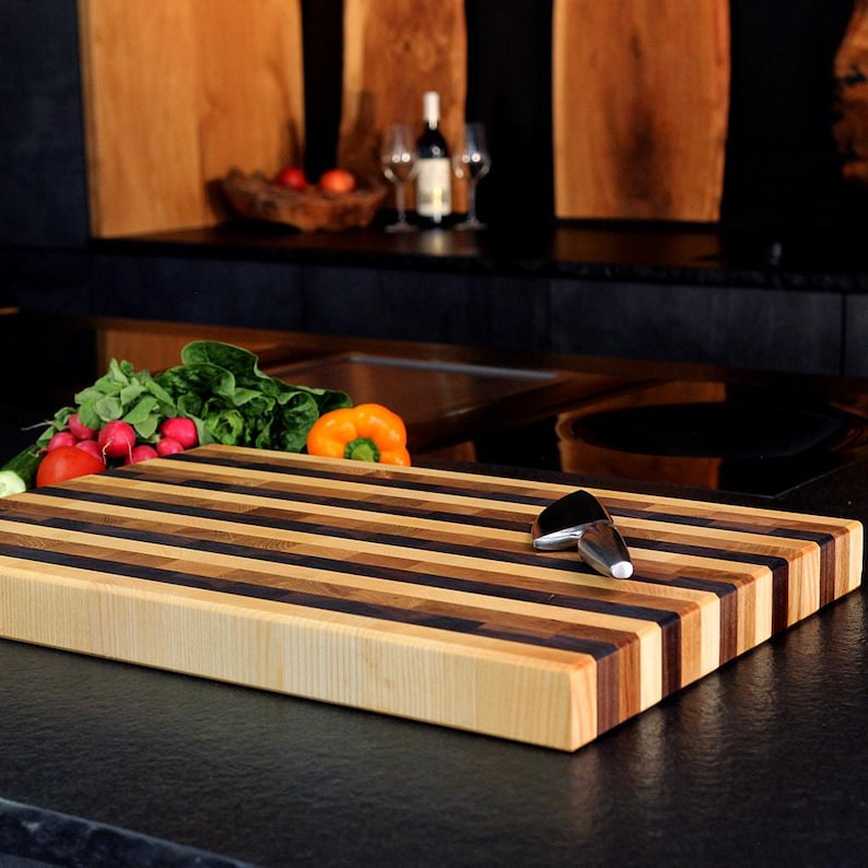 Peut inclure: Une planche &agrave; d&eacute;couper rectangulaire en bois pr&eacute;sentant un motif ray&eacute; de tons clairs, moyens et fonc&eacute;s. Un petit couteau en m&eacute;tal repose sur la surface de la planche, plac&eacute;e pr&egrave;s d'un tas de laitue verte fra&icirc;che, de radis et d'un poivron orange.
