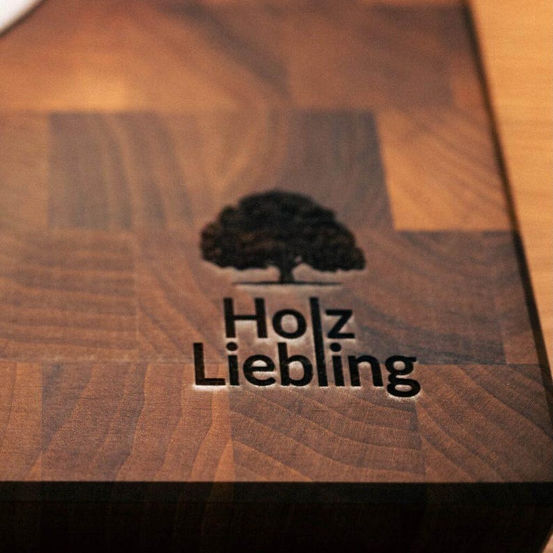 May include: A dark brown wooden chopping board with a visible end-grain pattern. The surface features a laser-engraved logo consisting of a stylised tree icon above the text "Holz Liebling" in a clean, sans-serif font centred on the board.