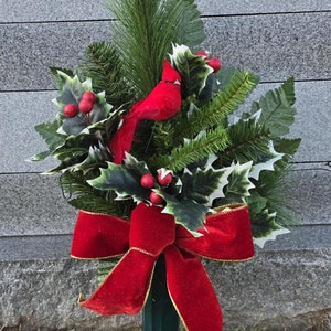 May include: A Christmas memorial arrangement featuring a vibrant red cardinal perched among holly leaves, berries, and evergreen branches. A large red velvet bow with gold trim adds a festive touch. The arrangement is set against a gray stone background.