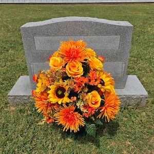 May include: A gray granite headstone with a rectangular inset, adorned with a vibrant floral arrangement. The arrangement features sunflowers, roses, and chrysanthemums in shades of orange and yellow, set against green grass.