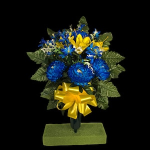 May include: A blue and yellow artificial flower arrangement with a yellow bow. The flowers are in a green vase and are sitting on a green base.