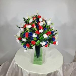 May include: A red, white, and blue artificial flower arrangement in a green vase. The arrangement is in a cemetery vase and is made of silk flowers.