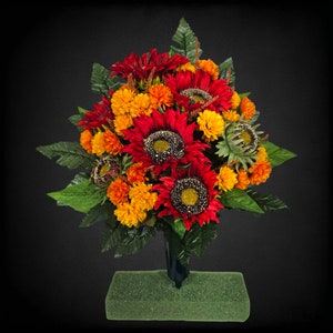 May include: A bouquet of artificial sunflowers and marigolds in shades of red, orange, and yellow. The flowers are arranged in a green foam base.