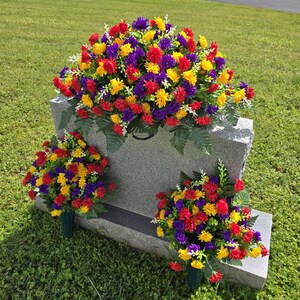 May include: A gray granite headstone adorned with vibrant, colorful floral arrangements. The flowers are a mix of red, yellow, and purple, with green foliage. The arrangement includes a large top piece and two smaller vase arrangements.