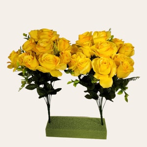 May include: Two bunches of yellow silk roses with green leaves arranged in a green floral foam block.