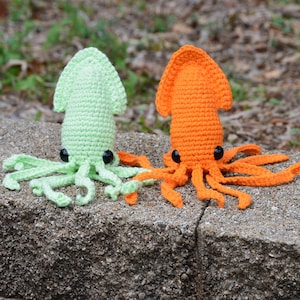 Puede incluir: Dos peluches de calamar tejidos a crochet, uno verde y otro naranja, con ojos de botón negros. Los peluches están sentados sobre una superficie de concreto gris.