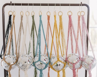 May include: A collection of disco ball plant hangers in various colors, including black, tan, cream, green, teal, orange, yellow, pink, and light pink. Each ball is suspended by a macrame hanger with a wooden ring.