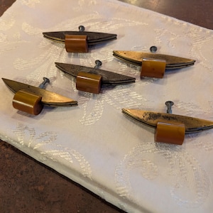 May include: Five vintage drawer pulls with a unique design. Each pull features a dark, wing-shaped metal plate with a screw and a cylindrical, amber-colored handle. The pulls are arranged on a white patterned fabric.
