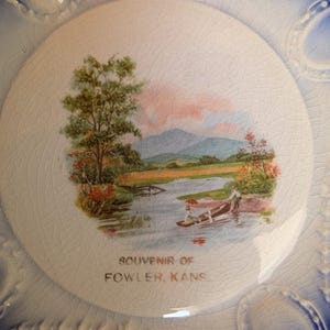 May include: A vintage ceramic plate with a painted landscape scene featuring a river, trees, and mountains. The plate has a white center with a cracked glaze and a blue border with decorative oval accents. The text "SOUVENIR OF FOWLER, KANS" is printed on the plate.
