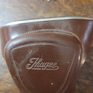 May include: A brown leather camera case with a silver trim and the word "Thayer" embossed on the front.