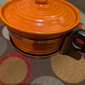 May include: A round orange ceramic casserole dish with a matching lid featuring a concentric circular pattern. The dish is encased in a metallic band and includes a dark brown handle attached to the side, providing a functional grip for the vessel.