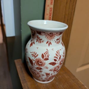 May include: A cream-colored ceramic vase featuring a hand-painted reddish-brown floral pattern. The vase is positioned next to a wooden ruler showing a height of approximately 6 inches. The vase has a flared rim and a rounded body.