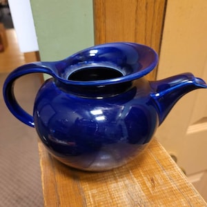 May include: A cobalt blue ceramic teapot with a wide, rounded body and a spout on the right side. The teapot has a handle on the left side and a wide, flat lid.