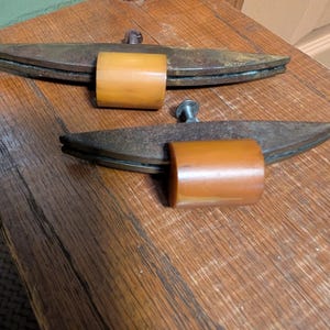 May include: Two drawer pulls featuring a long, curved, weathered metal bar with a dark, rusted finish. Each pull has a smooth, cylindrical, amber-colored center piece made of a solid material, with a single metal bolt extending from the back.