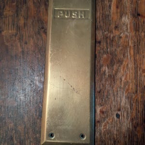 May include: A rectangular brass door push plate with a weathered, metallic finish. The plate features the word PUSH embossed in capital letters near the top and includes four small circular screw holes, two at the top and two at the bottom.