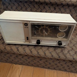 May include: A white rectangular Westinghouse FM/AM radio with an integrated analog clock face. The device features a left-side speaker grille, a central clock with hands, two circular tuning dials, and two black control knobs on the front panel.