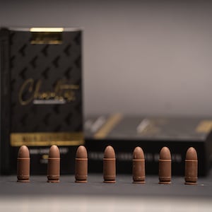 May include: A row of seven chocolate bullets are displayed in front of a black box with gold lettering that reads "Chocolate". The bullets are brown and have a realistic shape. The box has a repeating arrow pattern.