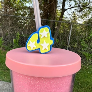 Può includere: Un bicchiere rosa con glitter con un topper a forma di numero quattro blu e giallo con stelle bianche. Una cannuccia di plastica trasparente è inserita nel topper.