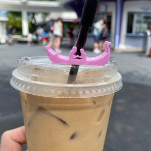 May include: A clear plastic cup filled with iced coffee and a black straw with a pink gondola-shaped straw topper.