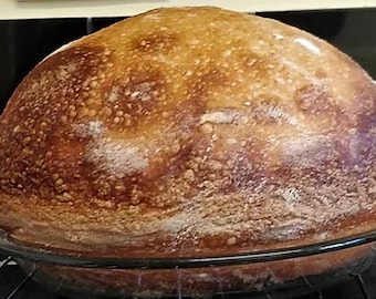 May include: A large, round loaf of bread with a golden brown crust and a slightly cracked surface. The bread is sitting in a clear glass baking dish on a wire rack.