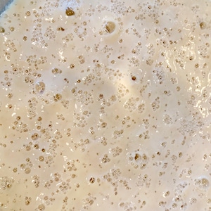 Pode incluir: Um close-up de uma mistura bege claro com muitas bolhas pequenas, indicando que está a crescer. Esta é uma visão comum ao fazer massa de pão.