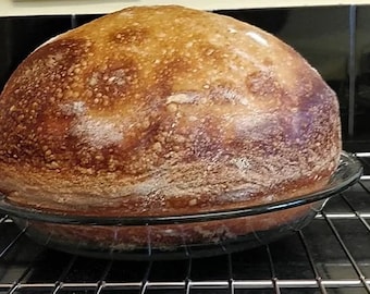 May include: A large, round loaf of bread with a golden brown crust and a slightly cracked surface. The bread is in a glass baking dish and is sitting on a wire rack.