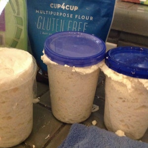 以下が含まれることがあります： 青い蓋が付いた3つの透明なプラスチック容器。中身は薄い色の物質で、おそらく生地またはバッターです。「Cup4Cup Multipurpose Flour Gluten Free」と書かれた青い袋が背景にあります。青い布が手前にあります。
