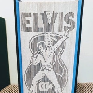 May include: A black and white silhouette of Elvis Presley singing into a microphone with a guitar behind him. The image is on a white background with the word "ELVIS" in black letters at the top. The image is framed with a black border and a blue border.