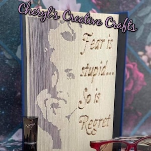 May include: A wooden book art piece with a woman's face carved into it. The text "Fear is stupid... So is Regret" is carved into the wood. The piece is sitting on a table with a lipstick and glasses.