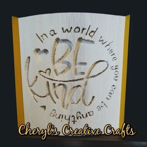 May include: A white paper book with a yellow border, folded into a 3D sculpture. The sculpture is a circle with the words "In a world where you can be anything, be kind" and a heart in the center. The words "Cheryl's Creative Crafts" are printed below the sculpture.