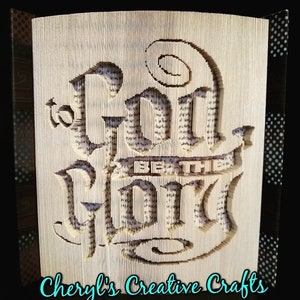 Puede incluir: Una escultura de arte de libro con las palabras "a Dios sea la gloria" dobladas en las páginas. La escultura está hecha de un libro con una cubierta marrón. Las palabras están en una fuente decorativa y se resaltan en blanco. La escultura está sobre un fondo con patrones en blanco y negro. Las palabras "Cheryl's Creative Crafts" están escritas en turquesa debajo de la escultura.