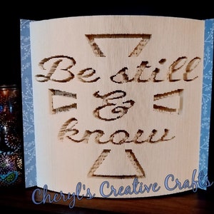 Könnte beinhalten: Ein beigefarbenes Papierhandwerk mit den Worten "Be still & know" in einer dekorativen Schriftart. Das Papierhandwerk ist zu einer dreidimensionalen Form gefaltet und hat einen blau-weißen Musterrand. Die Worte "Cheryl's Creative Crafts" sind unten aufgedruckt.