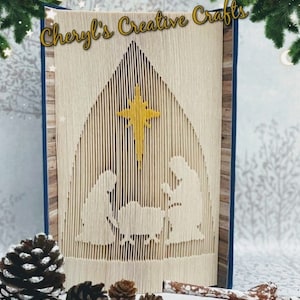 May include: A folded book art piece featuring a nativity scene with a gold star. The book is beige with a blue spine and the text "Cheryl's Creative Crafts" in gold. Pinecones and greenery frame the scene.