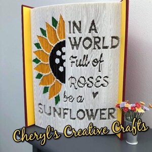 Peut inclure: Une œuvre d'art de livre avec un motif de tournesol et le texte "Dans un monde plein de roses, soyez un tournesol". Le livre est ouvert et posé sur une surface grise. Un petit vase avec des fleurs colorées se trouve à droite de l'image. Le texte "Cheryl's Creative Crafts" est en bas de l'image.
